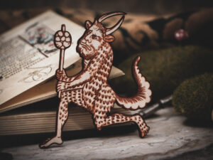 A laser cut and engraved magnet made from raw birch plywood in the shape of a satyr holding a branch from the Aberdeen Bestiary leans against an open illuminated manuscript resting on a stone surface surrounded by mossy stones.