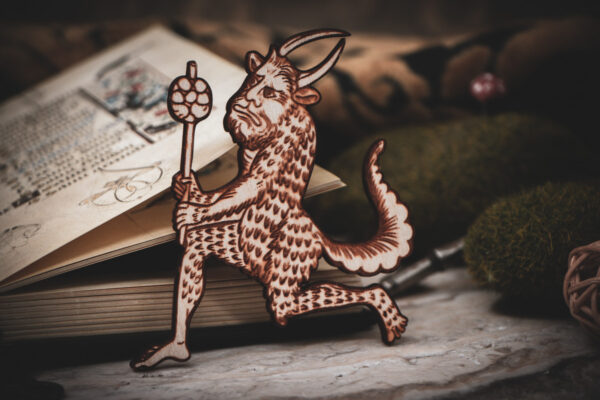 A laser cut and engraved magnet made from raw birch plywood in the shape of a satyr holding a branch from the Aberdeen Bestiary leans against an open illuminated manuscript resting on a stone surface surrounded by mossy stones.