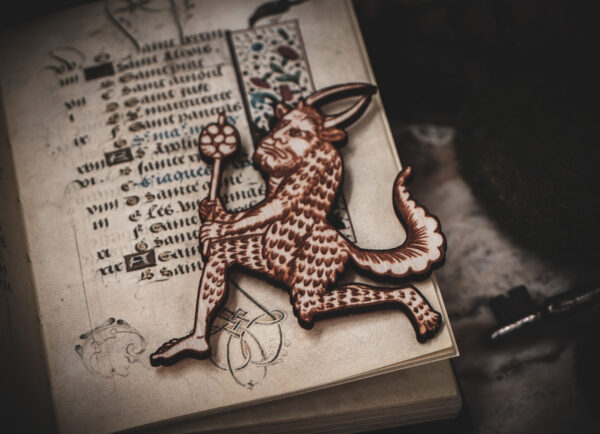 A laser cut and engraved magnet made from raw birch plywood in the shape of a satyr holding a branch from the Aberdeen Bestiary on top of the page of an illuminated manuscript.