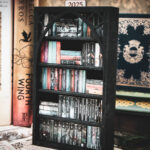A laser cut mini bookshelf with a gothic arch detail with two quatrefoils in the top right corners above the arch filled with mini 3D printed books with vinyl sticker covers in front of full-size books on a stone surface.