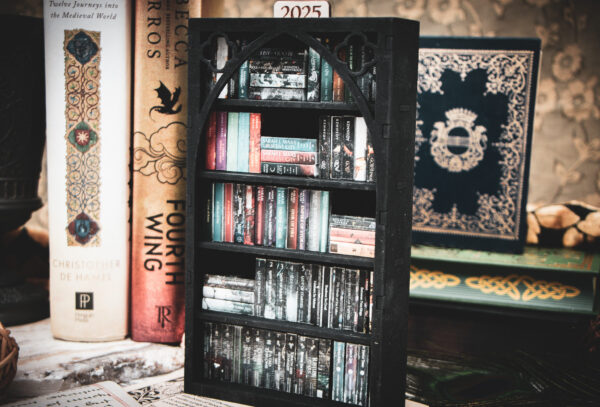 A laser cut mini bookshelf with a gothic arch detail with two quatrefoils in the top right corners above the arch filled with mini 3D printed books with vinyl sticker covers in front of full-size books on a stone surface.