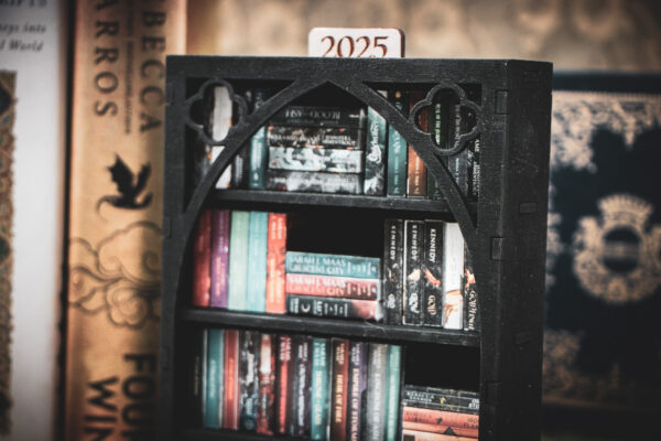 Detail of the top of a laser cut mini bookshelf with a gothic arch detail with two quatrefoils in the top right corners above the arch filled with mini 3D printed books with vinyl sticker covers in front of full-size books on a stone surface.