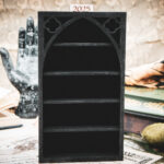 A tall, laser-cut wooden miniature bookshelf in black, featuring a prominent gothic arch and intricate quatrefoil cutouts at the top.