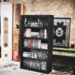 A laser cut mini bookshelf with a gothic arch detail with two quatrefoils in the top right corners above the arch filled with mini 3D printed books with vinyl sticker covers in front of full-size books on a stone surface.