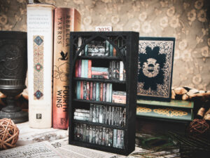 A laser cut mini bookshelf with a gothic arch detail with two quatrefoils in the top right corners above the arch filled with mini 3D printed books with vinyl sticker covers in front of full-size books on a stone surface.