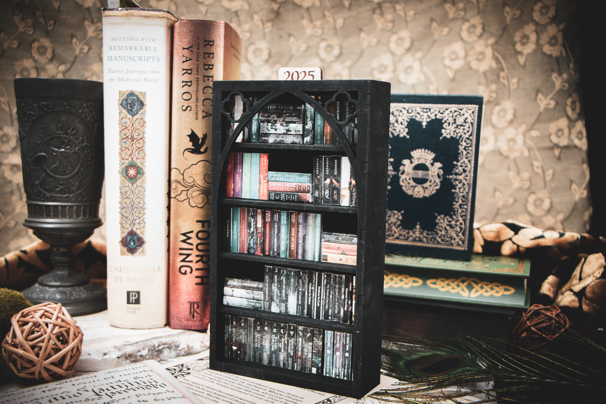 The Cathedral Archivist DIY Library - The Griffin and Stag A laser cut mini bookshelf with a gothic arch detail with two quatrefoils in the top right corners above the arch filled with mini 3D printed books with vinyl sticker covers in front of full-size books on a stone surface.