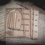 The pieces of a A laser cut mini bookshelf with a gothic arch detail with two quatrefoils in the top right corners above the arch arranged in a group on top of various papers