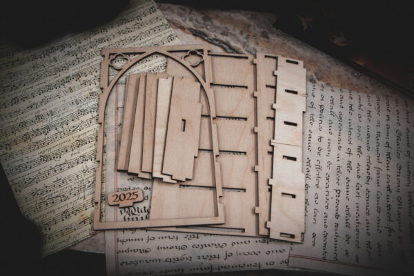 The pieces of a A laser cut mini bookshelf with a gothic arch detail with two quatrefoils in the top right corners above the arch arranged in a group on top of various papers
