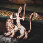 A laser-cut wooden magnet of a medieval-style rabbit knight holding a sword and shield while riding a lion, displayed against dark green hardcover books with gold Celtic knots.