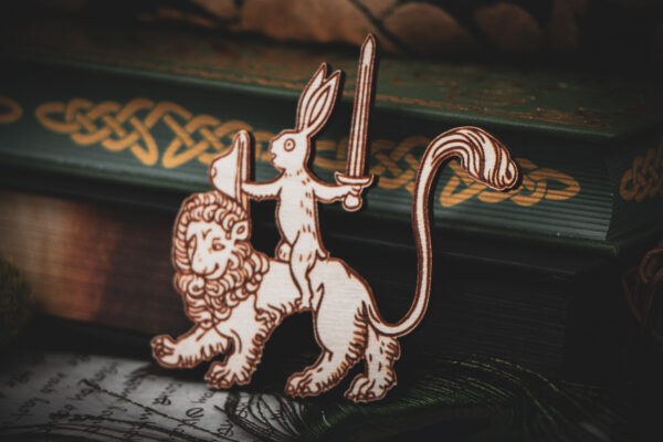 A laser-cut wooden magnet of a medieval-style rabbit knight holding a sword and shield while riding a lion, displayed against dark green hardcover books with gold Celtic knots.