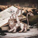 Handcrafted birch plywood magnet featuring a whimsical medieval "drollery" illustration of a rabbit riding a lion, styled on a marble surface next to an open antique manuscript and mossy stones.