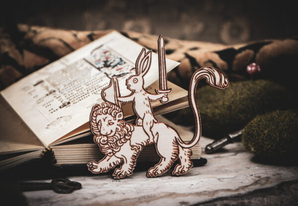Handcrafted birch plywood magnet featuring a whimsical medieval "drollery" illustration of a rabbit riding a lion, styled on a marble surface next to an open antique manuscript and mossy stones.