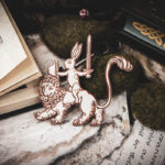 Handcrafted birch plywood magnet featuring a whimsical medieval "drollery" illustration of a rabbit riding a lion, styled on a marble surface next to an open antique manuscript and mossy stones.