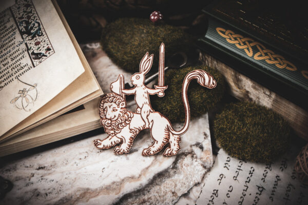 Handcrafted birch plywood magnet featuring a whimsical medieval "drollery" illustration of a rabbit riding a lion, styled on a marble surface next to an open antique manuscript and mossy stones.