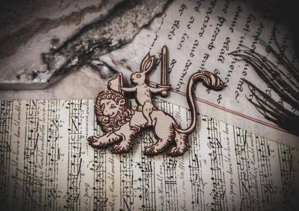 Close-up of a wooden laser-engraved magnet of a rabbit knight on a lion, placed over vintage sheet music and handwritten calligraphy.