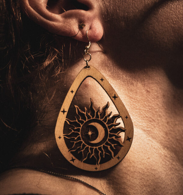 A laser cut and engraved earring featuring a sun with a crescent moon and eight-pointed start in its center within a teardrop frame hanging from the ear of a model.
