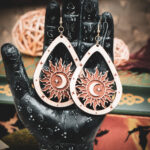 Laser cut and engraved earrings featuring a sun with a crescent moon and eight-pointed start in its center within a teardrop frame hanging from the fingers of a hand sculpture.