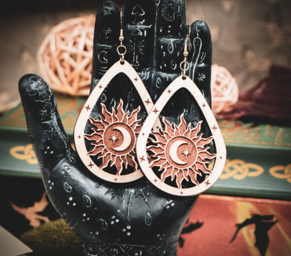 Laser cut and engraved earrings featuring a sun with a crescent moon and eight-pointed start in its center within a teardrop frame hanging from the fingers of a hand sculpture.