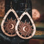 Laser cut and engraved earrings featuring a sun with a crescent moon and eight-pointed start in its center within a teardrop frame hanging from the fingers of a hand sculpture.