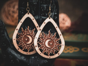 Laser cut and engraved earrings featuring a sun with a crescent moon and eight-pointed start in its center within a teardrop frame hanging from the fingers of a hand sculpture.