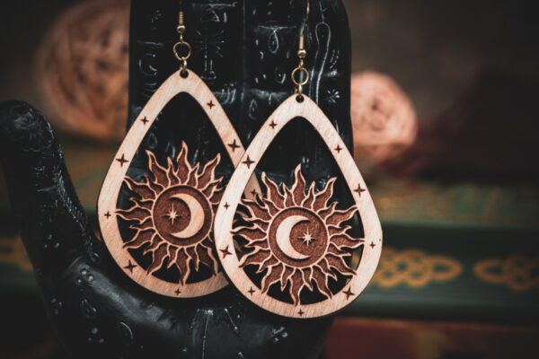 Laser cut and engraved earrings featuring a sun with a crescent moon and eight-pointed start in its center within a teardrop frame hanging from the fingers of a hand sculpture.