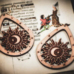 Handcrafted birch wood celestial earrings with sun and moon engravings, styled atop an open medieval manuscript featuring whimsical drollery illustrations.