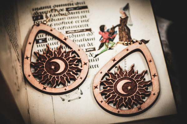 Handcrafted birch wood celestial earrings with sun and moon engravings, styled atop an open medieval manuscript featuring whimsical drollery illustrations.