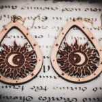 Laser cut and engraved earrings featuring a sun with a crescent moon and eight-pointed start in its center within a teardrop frame on top of a page of calligraphy.