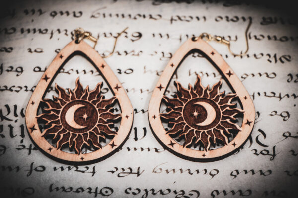 Laser cut and engraved earrings featuring a sun with a crescent moon and eight-pointed start in its center within a teardrop frame on top of a page of calligraphy.