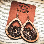 Laser cut and engraved earrings featuring a sun with a crescent moon and eight-pointed start in its center within a teardrop frame hanging from a kraft paper card on top of the page of an open book.