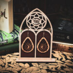 A pair of teardrop-shaped wooden earrings, the "Celestial Alignment," featuring a laser-cut sun, crescent moon, and star, displayed on a custom gothic-arch stand with The Griffin and Stag logo.