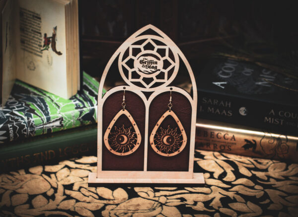 A pair of teardrop-shaped wooden earrings, the "Celestial Alignment," featuring a laser-cut sun, crescent moon, and star, displayed on a custom gothic-arch stand with The Griffin and Stag logo.
