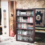 A laser cut mini bookshelf filled with mini 3D printed books with vinyl sticker covers in front of some full-size books on a stone surface at a slight angle