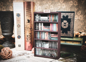 A laser cut mini bookshelf filled with mini 3D printed books with vinyl sticker covers in front of some full-size books on a stone surface at a slight angle