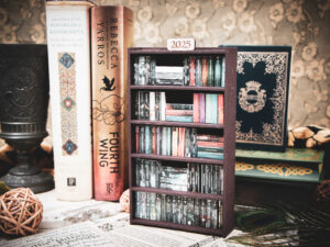 A laser cut mini bookshelf filled with mini 3D printed books with vinyl sticker covers in front of some full-size books on a stone surface at a slight angle