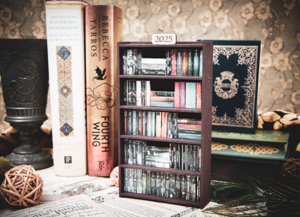 A laser cut mini bookshelf filled with mini 3D printed books with vinyl sticker covers in front of some full-size books on a stone surface at a slight angle