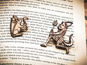 A set of two pins, one featuring a rabbit with a shield on its back holding an axe, the other featuring a medieval satyr holding a branch, laser engraved on raw birch on top of the page of a book.