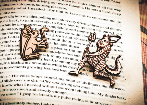 A set of two pins, one featuring a rabbit with a shield on its back holding an axe, the other featuring a medieval satyr holding a branch, laser engraved on raw birch on top of the page of a book.