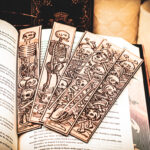 Five laser cut and engraved bookmarks featuring various scenes of skeletons and bones in a medieval woodcut style on the page of an open book.