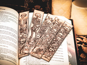 Five laser cut and engraved bookmarks featuring various scenes of skeletons and bones in a medieval woodcut style on the page of an open book.