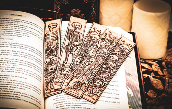 Five laser cut and engraved bookmarks featuring various scenes of skeletons and bones in a medieval woodcut style on the page of an open book.