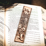 A laser cut and engraved bookmark featuring a scene of skeletons and bones in a medieval woodcut style on the page of an open book.
