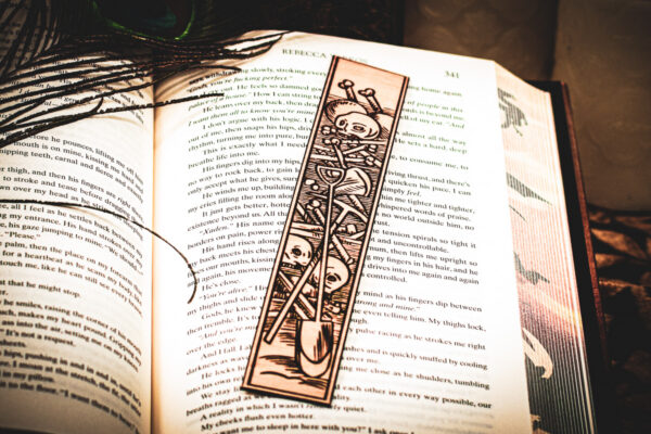 A laser cut and engraved bookmark featuring a scene of skeletons and bones in a medieval woodcut style on the page of an open book.