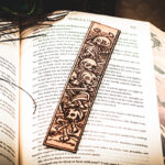 A laser cut and engraved bookmark featuring a scene of skeletons and bones in a medieval woodcut style on the page of an open book.