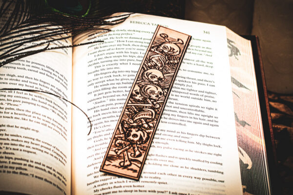 A laser cut and engraved bookmark featuring a scene of skeletons and bones in a medieval woodcut style on the page of an open book.