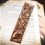 A laser cut and engraved bookmark featuring a scene of skeletons and bones in a medieval woodcut style on the page of an open book.
