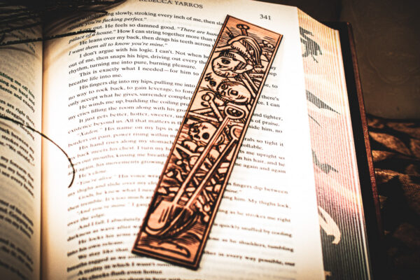 A laser cut and engraved bookmark featuring a scene of skeletons and bones in a medieval woodcut style on the page of an open book.