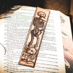 A laser cut and engraved bookmark featuring a scene of skeletons and bones in a medieval woodcut style on the page of an open book.