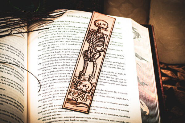 A laser cut and engraved bookmark featuring a scene of skeletons and bones in a medieval woodcut style on the page of an open book.