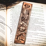 A laser cut and engraved bookmark featuring a scene of skeletons and bones in a medieval woodcut style on the page of an open book.